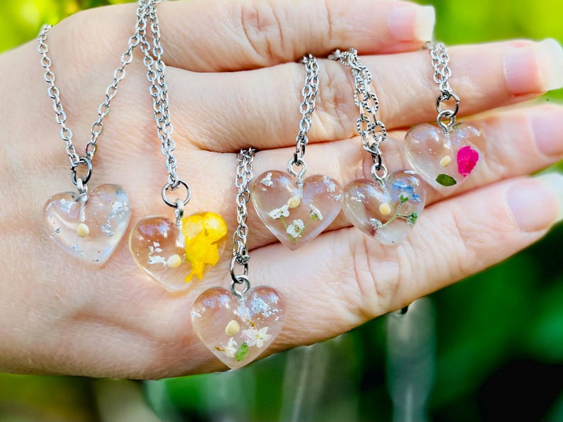 Puede incluir: Cinco collares de plata con colgantes en forma de coraz&oacute;n rellenos de flores prensadas. Cada colgante presenta una disposici&oacute;n diferente de flores, incluidas flores blancas, amarillas, rosas y azules.