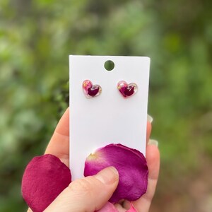 May include: A pair of heart-shaped earrings with pink and purple pressed flowers inside. The earrings are on a white card with a pink rose petal in the foreground.