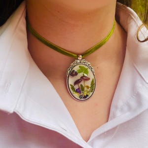 K&ouml;nnte beinhalten: Eine gr&uuml;ne Samt-Chokerkette mit einem silbernen ovalen Anh&auml;nger mit einem gepressten Blumen- und Pilzdesign.