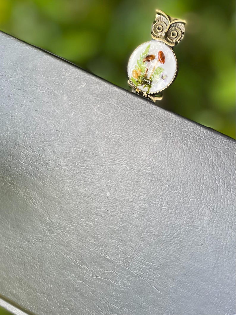 May include: A gold owl bookmark with a white background and a mushroom design. The owl is perched on a grey leather surface.
