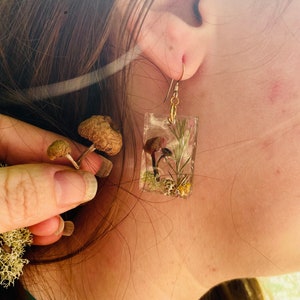 May include: A pair of clear resin earrings with a gold hook. The earrings feature a small mushroom and green foliage encased in the resin.