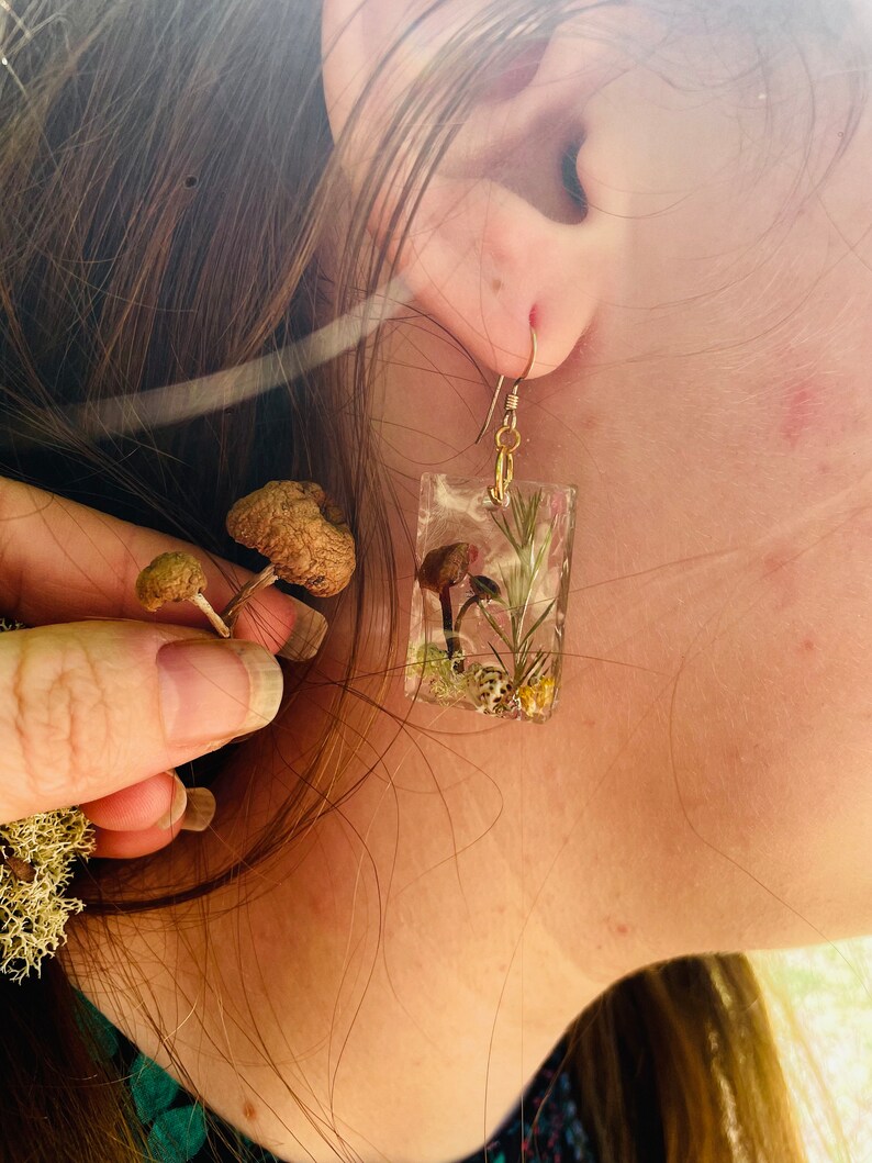 May include: A pair of clear resin earrings with a gold hook. The earrings feature a small mushroom and green foliage encased in the resin.