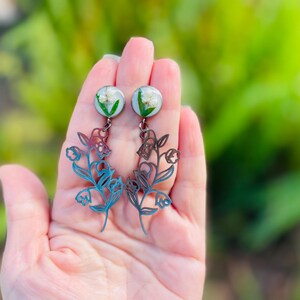 May include: A pair of dangle earrings featuring a floral design. The earrings have a round, clear top with white flowers and green accents. The lower portion is a blue and brown floral cutout. The earrings are held in a hand against a blurred green background.