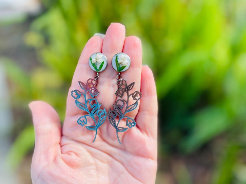 May include: A pair of dangle earrings featuring a floral design. The earrings have a round, clear top with white flowers and green accents. The lower portion is a blue and brown floral cutout. The earrings are held in a hand against a blurred green background.