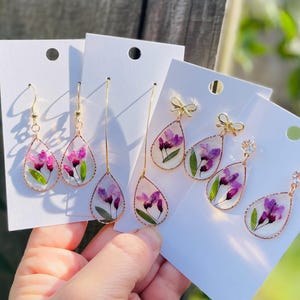 Puede incluir: Pendientes dorados en forma de lágrima con flores moradas prensadas y hojas verdes encapsuladas en resina transparente. Algunos pendientes presentan lazos dorados o un pequeño cristal. Los pendientes se exhiben en tarjetas blancas.
