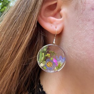 Puede incluir: Pendientes circulares transparentes con flores secas encapsuladas en resina. Los pendientes presentan una variedad de flores coloridas, incluyendo flores moradas, azules y amarillas, y follaje verde. Cuelgan de ganchos plateados.