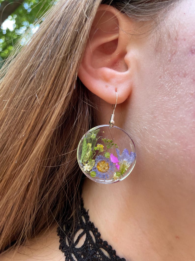 Puede incluir: Pendientes circulares transparentes con flores secas encapsuladas en resina. Los pendientes presentan una variedad de flores coloridas, incluyendo flores moradas, azules y amarillas, y follaje verde. Cuelgan de ganchos plateados.