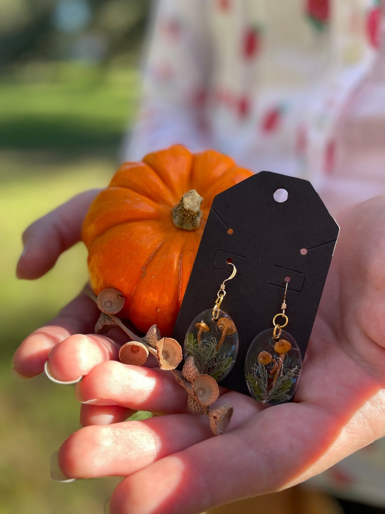 May include: A pair of gold earrings with oval-shaped resin pendants featuring pressed mushrooms and greenery. The earrings are displayed on a black card with a pumpkin in the background.