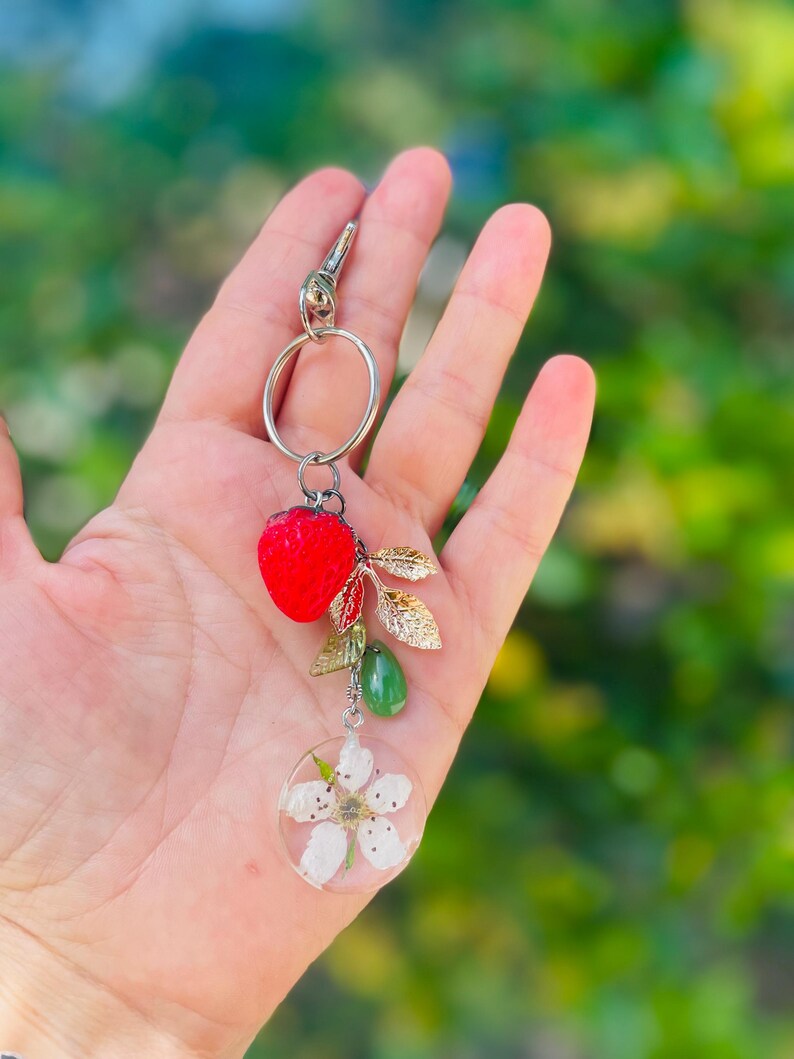 Peut inclure: Un porte-cl&eacute;s argent&eacute; avec un charm en forme de fraise rouge, des feuilles dor&eacute;es, une goutte verte et un pendentif circulaire transparent avec une fleur blanche. Le porte-cl&eacute;s est tenu sur un fond vert flou.