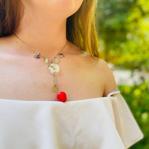 May include: A silver necklace with a white flower charm and a red heart pendant. The necklace is made with green and purple stones.