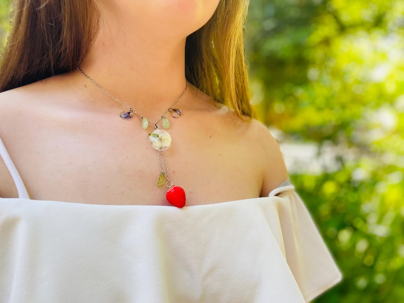 May include: A silver necklace with a white flower charm and a red heart pendant. The necklace is made with green and purple stones.