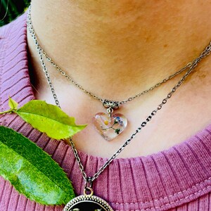 Puede incluir: Un collar de cadena de plata con un colgante en forma de coraz&oacute;n. El colgante est&aacute; hecho de resina transparente y contiene flores prensadas. El collar est&aacute; superpuesto a una segunda cadena de plata.