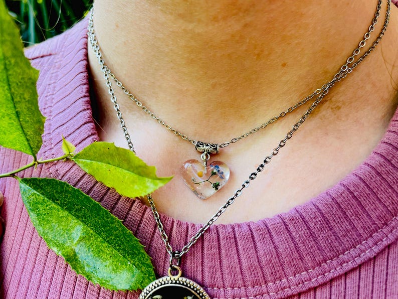 Puede incluir: Un collar de cadena de plata con un colgante en forma de coraz&oacute;n. El colgante est&aacute; hecho de resina transparente y contiene flores prensadas. El collar est&aacute; superpuesto a una segunda cadena de plata.