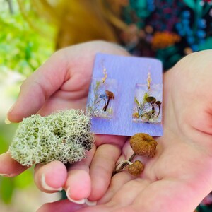 May include: A pair of resin earrings with dried flowers and mushrooms. The earrings are on a purple card. There are also dried mushrooms and moss in the image.