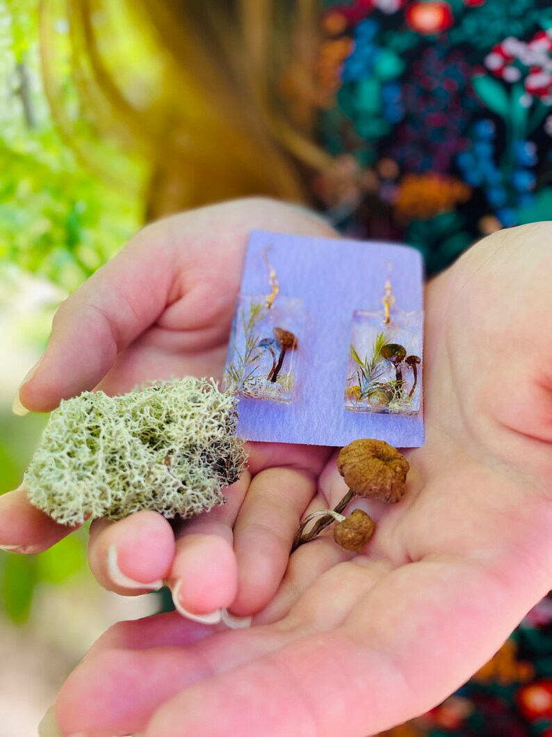 May include: A pair of resin earrings with dried flowers and mushrooms. The earrings are on a purple card. There are also dried mushrooms and moss in the image.