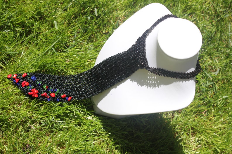 May include: A black beaded necklace with a decorative end featuring red, blue, and green accents. The necklace is displayed on a white mannequin head against a backdrop of green grass.