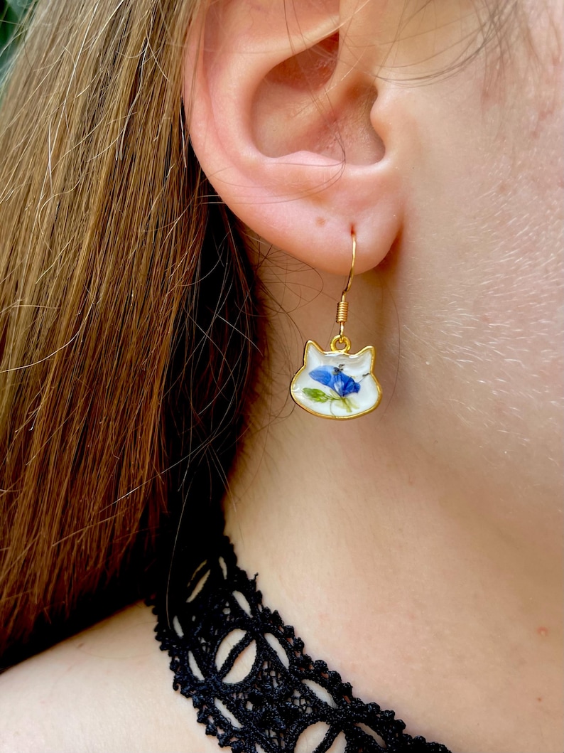 Puede incluir: Pendientes dorados con forma de gato y acabado esmaltado blanco. Cada pendiente presenta un dise&ntilde;o de flor azul. Los pendientes est&aacute;n suspendidos de ganchos dorados. Un accesorio &uacute;nico.