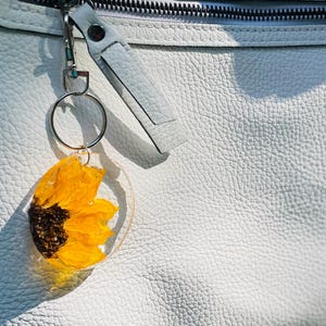 May include: A clear acrylic keyring featuring a pressed yellow sunflower with a brown centre. The keyring is attached to a silver ring and a white leather strap, set against a textured white leather background.