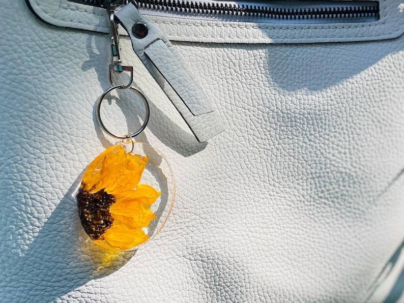 May include: A clear acrylic keyring featuring a pressed yellow sunflower with a brown centre. The keyring is attached to a silver ring and a white leather strap, set against a textured white leather background.