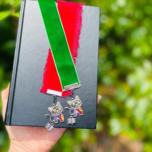 May include: Two velvet bookmarks with fringe, one red and one green, attached to silver cat charms. The charms have black and white eyes and a colorful design.