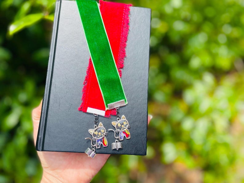 May include: Two velvet bookmarks with fringe, one red and one green, attached to silver cat charms. The charms have black and white eyes and a colorful design.