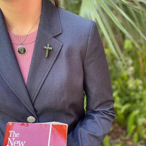 May include: A person wearing a dark blue blazer with a silver cross pin and a silver necklace with a round pendant. The person is holding a red book with the title "The New American Bible".