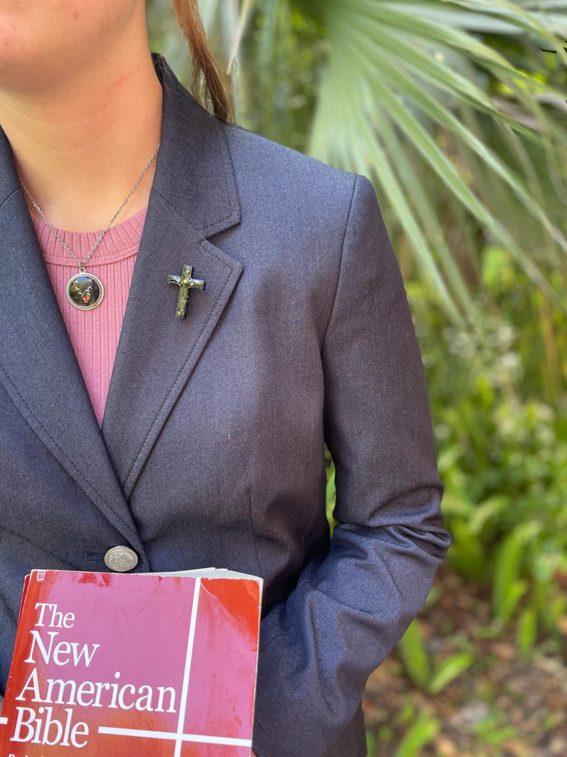 May include: A person wearing a dark blue blazer with a silver cross pin and a silver necklace with a round pendant. The person is holding a red book with the title "The New American Bible".