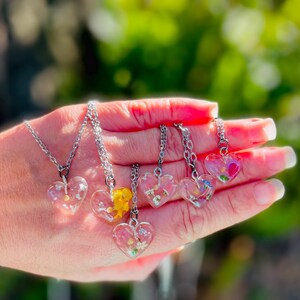 Puede incluir: Seis collares de plata con colgantes de coraz&oacute;n transparentes llenos de flores prensadas. Los colgantes est&aacute;n llenos de flores de diferentes colores, incluyendo rosa, amarillo y blanco.