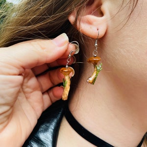 May include: Mushroom-shaped earrings with a brown cap and a beige stem, accented with green foliage. The earrings hang from silver-colored hooks. The earrings are a unique, nature-inspired accessory.