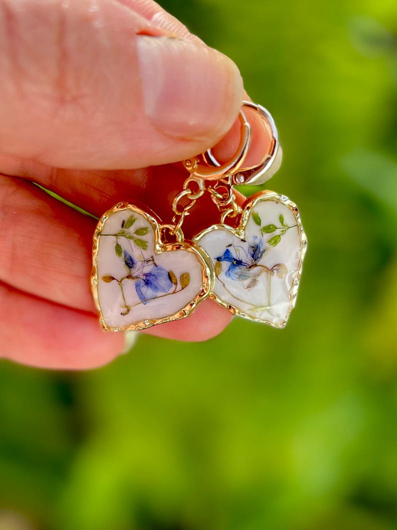 May include: A pair of gold heart-shaped earrings with pressed flowers inside. The flowers are blue, green, and white.