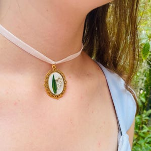 Pode incluir: Um colar de choker de cetim branco com um pingente oval dourado que apresenta uma flor de l&iacute;rio do vale prensada.