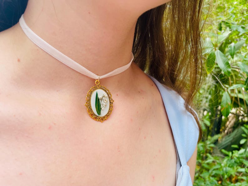 Pode incluir: Um colar de choker de cetim branco com um pingente oval dourado que apresenta uma flor de l&iacute;rio do vale prensada.