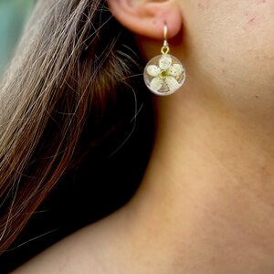 Puede incluir: Un par de pendientes de oro con un colgante de resina transparente que contiene una flor blanca prensada.