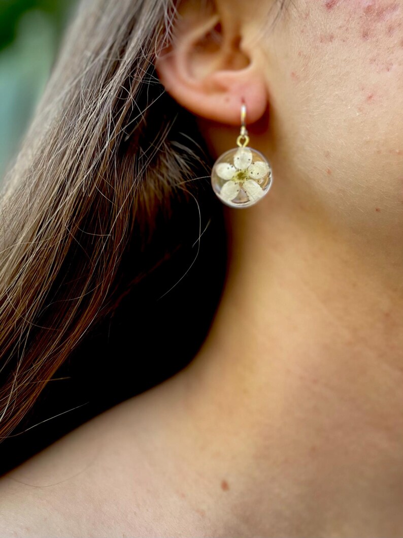 Puede incluir: Un par de pendientes de oro con un colgante de resina transparente que contiene una flor blanca prensada.