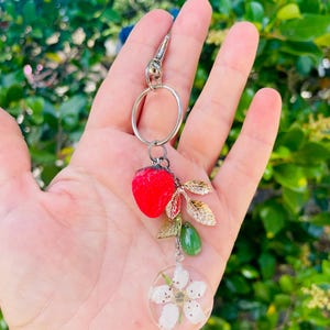 Peut inclure: Un porte-cl&eacute;s argent&eacute; avec un charm en forme de fraise rouge, des feuilles dor&eacute;es, une perle verte en forme de goutte et un pendentif circulaire transparent avec des fleurs blanches. Le porte-cl&eacute;s est tenu dans une main sur un fond vert flou.