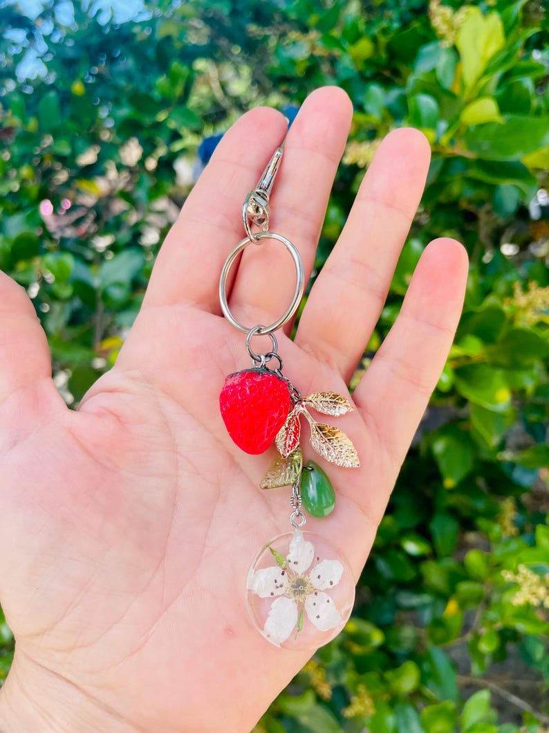 Peut inclure: Un porte-cl&eacute;s argent&eacute; avec un charm en forme de fraise rouge, des feuilles dor&eacute;es, une perle verte en forme de goutte et un pendentif circulaire transparent avec des fleurs blanches. Le porte-cl&eacute;s est tenu dans une main sur un fond vert flou.