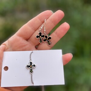May include: A set of silver cross earrings and a silver cross necklace with black enamel and gold accents. The earrings are dangling and the necklace has a chain.