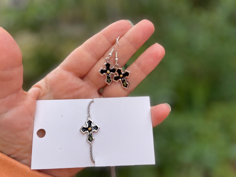 May include: A set of silver cross earrings and a silver cross necklace with black enamel and gold accents. The earrings are dangling and the necklace has a chain.