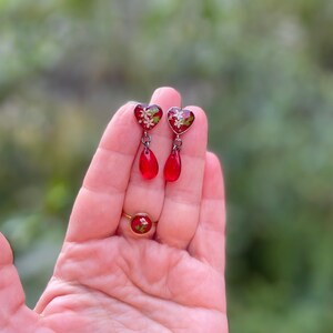 May include: A pair of red heart-shaped dangle earrings with a floral design and a teardrop-shaped dangle. The earrings are made of glass and have a silver-tone finish.