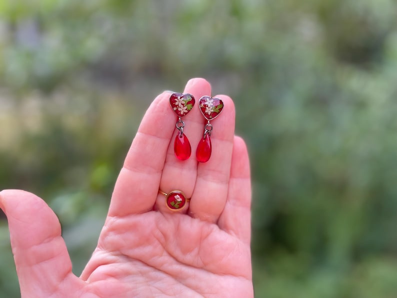 May include: A pair of red heart-shaped dangle earrings with a floral design and a teardrop-shaped dangle. The earrings are made of glass and have a silver-tone finish.
