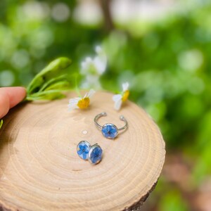 K&ouml;nnte beinhalten: Ein Set aus zwei Ringen mit blauen Blumenakzenten. Die Ringe sind aus Silber und haben ein schlichtes Design. Die Blumen sind gepresst und in Harz konserviert.