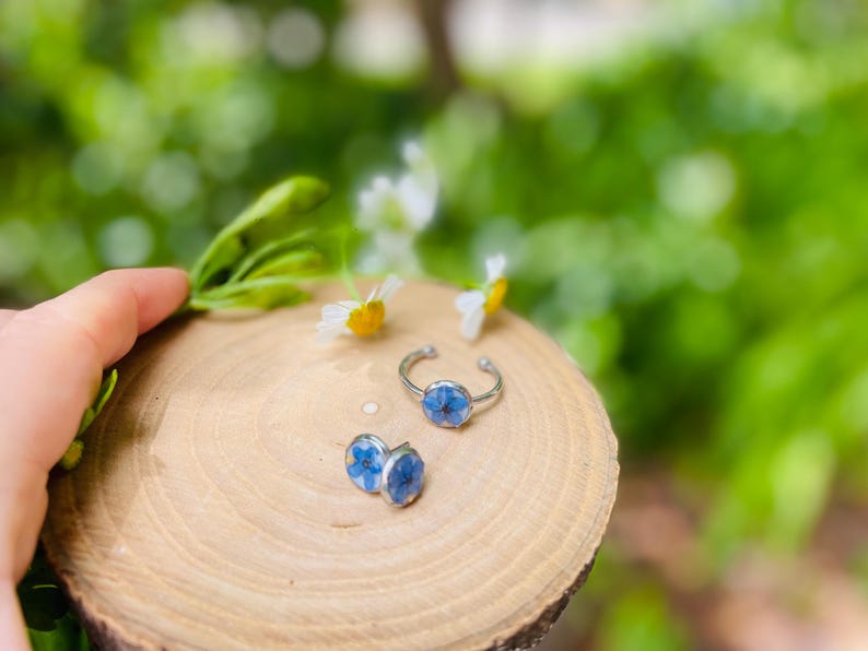 K&ouml;nnte beinhalten: Ein Set aus zwei Ringen mit blauen Blumenakzenten. Die Ringe sind aus Silber und haben ein schlichtes Design. Die Blumen sind gepresst und in Harz konserviert.