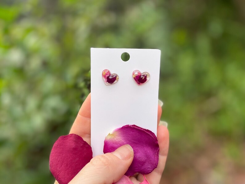 May include: A pair of heart-shaped earrings with pink and purple pressed flowers inside. The earrings are on a white card with a pink rose petal in the foreground.