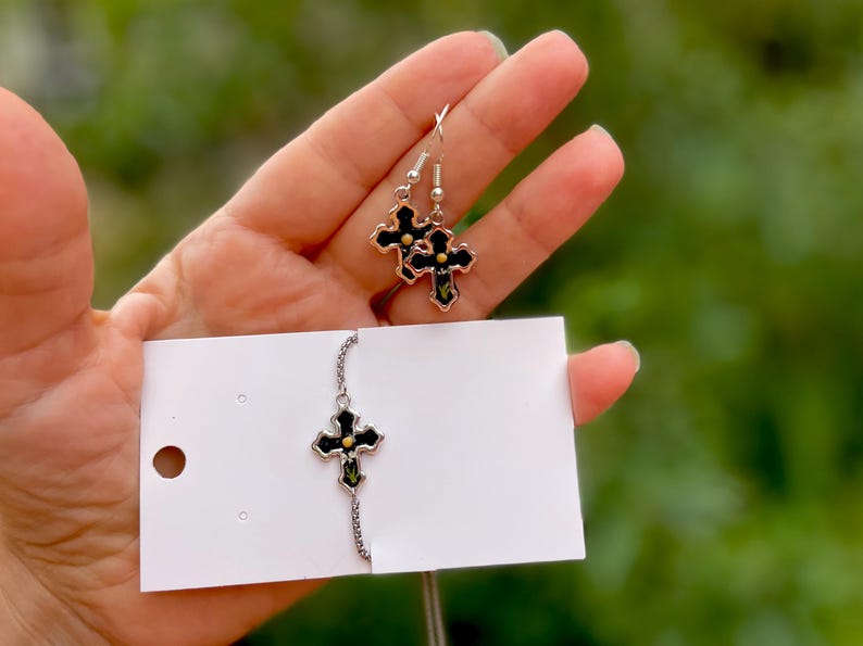 May include: A set of silver cross earrings and a silver cross necklace with black enamel and gold accents. The earrings and necklace feature a floral design.