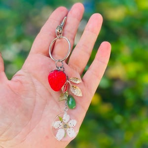 Peut inclure: Un porte-cl&eacute;s argent&eacute; avec un charm en forme de fraise rouge, des feuilles dor&eacute;es, une goutte verte et un pendentif circulaire transparent avec une fleur blanche. Le porte-cl&eacute;s est tenu sur un fond vert flou.