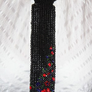 May include: A black beaded tie with a floral design. The tie is made of small black beads and features a pattern of red, blue, and green stones near the bottom. The tie is worn over a white collared shirt.