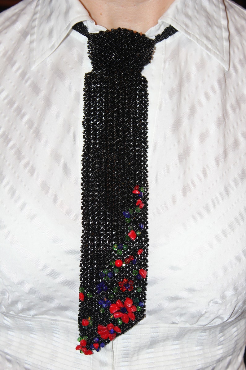 May include: A black beaded tie with a floral design. The tie is made of small black beads and features a pattern of red, blue, and green stones near the bottom. The tie is worn over a white collared shirt.
