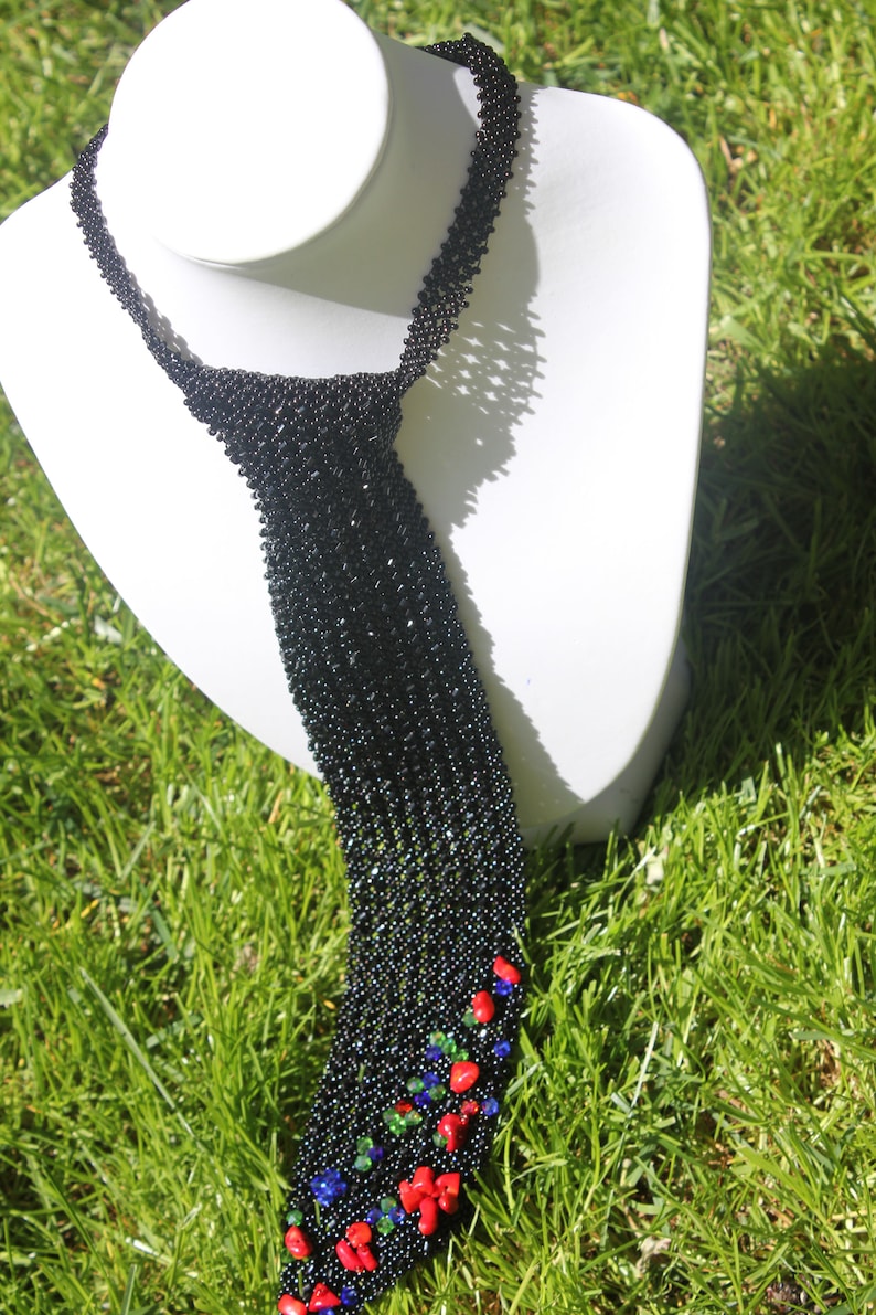 May include: A black beaded necklace with a tie-like design, displayed on a white mannequin. The bottom of the necklace features red, blue, and green accents. The necklace is set against a backdrop of green grass.