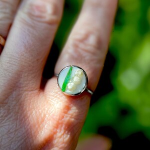 May include: A silver ring with a white and green resin inlay featuring a cluster of white flowers. The ring is on a finger.
