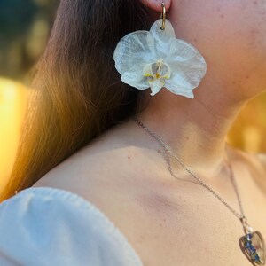 Pode incluir: Um brinco grande em forma de flor de orqu&iacute;dea branca transl&uacute;cida com um centro amarelo e roxo, suspenso por um aro fino de tom dourado. O brinco apresenta texturas de p&eacute;talas delicadas e realistas, exibindo um design bot&acirc;nico.